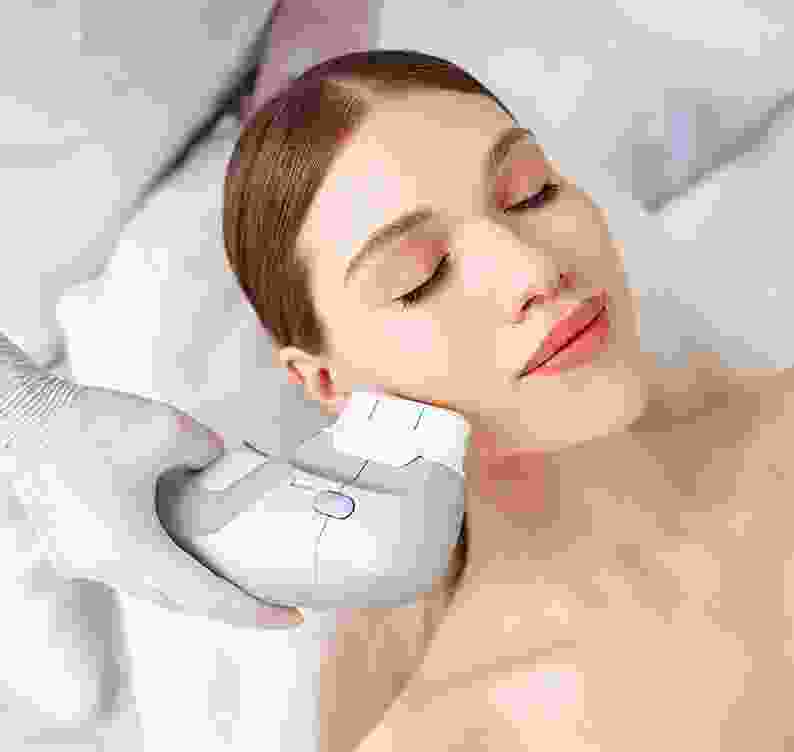 Woman reclining while a gloved practitioner holds a treatment handpiece against her cheek