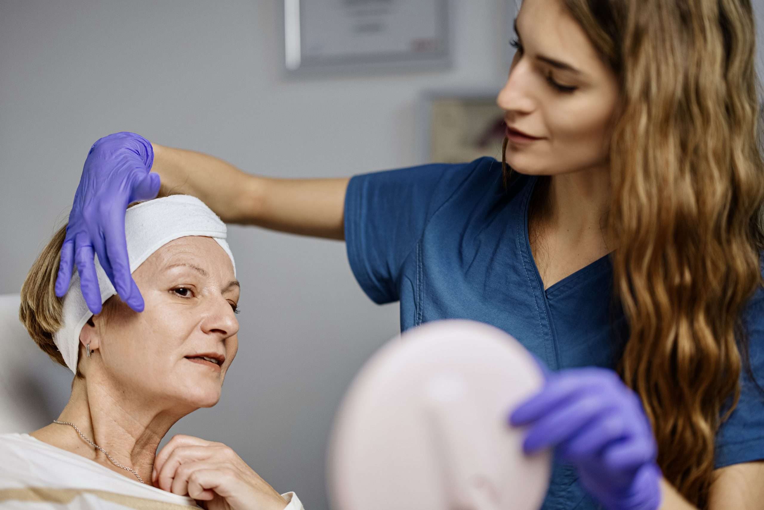 Practitioner wearing gloves discussing facial skin with a woman holding a mirror
